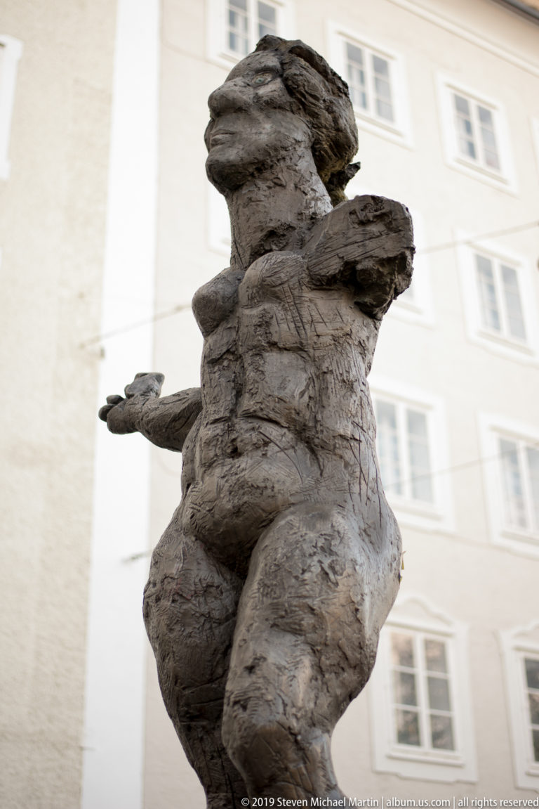 Close view of a weathered winged putto with outstretched arm against a softly blurred pale building.
