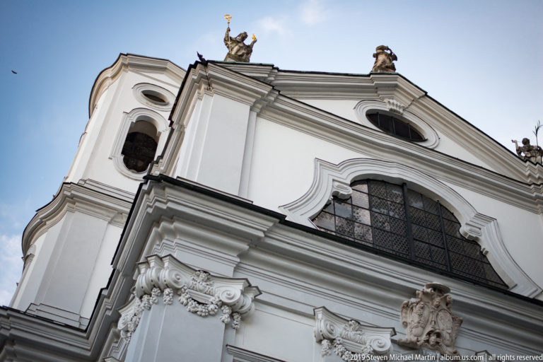 Upward view of a white Baroque church façade with arched windows, curved gables, pilasters, and sculptural figures.