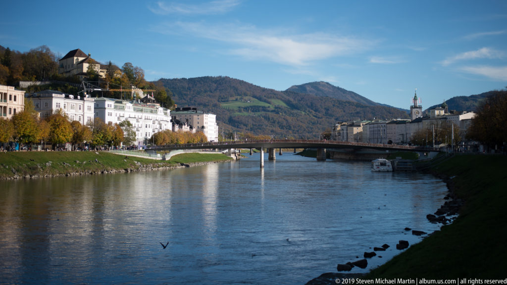 Wide view along the Salzach with a modern bridge, riverside buildings, wooded hills, and church towers in the distance.