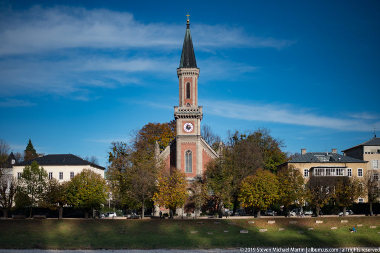 Red Gothic Revival church tower with pointed dark spire and clock, seen across a grassy bank beside the Salzach.