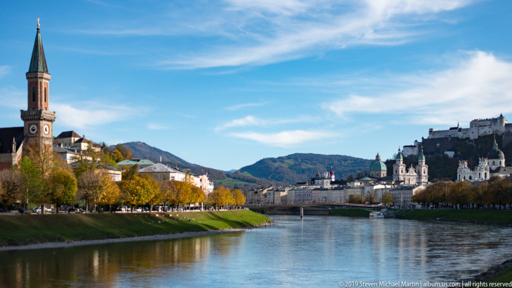 Salzach River with autumn-lined banks, a church tower at left, and Hohensalzburg Fortress beyond the historic center.