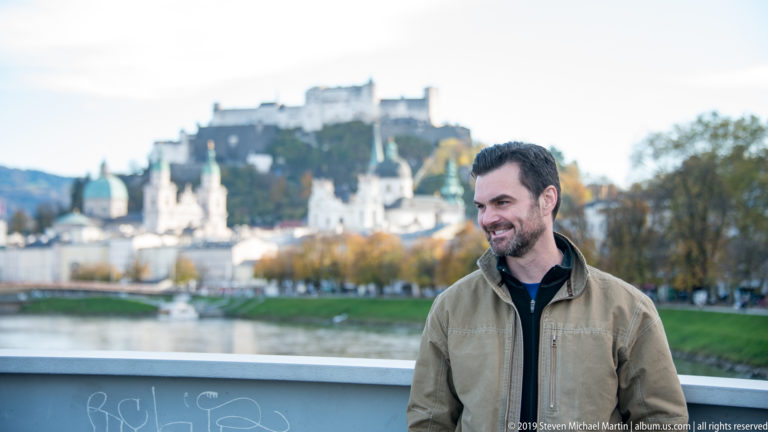 Smiling man in a tan jacket beside the Salzach, with blurred Hohensalzburg Fortress and old-city towers behind.
