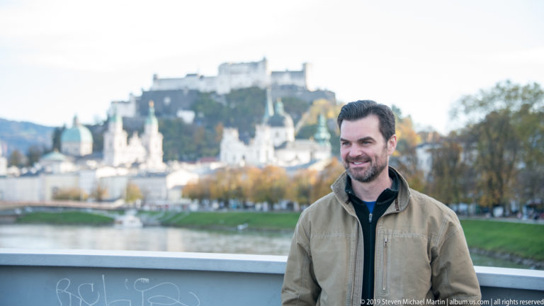 Smiling man beside the Salzach, with blurred church towers and Hohensalzburg Fortress on the hill behind him.