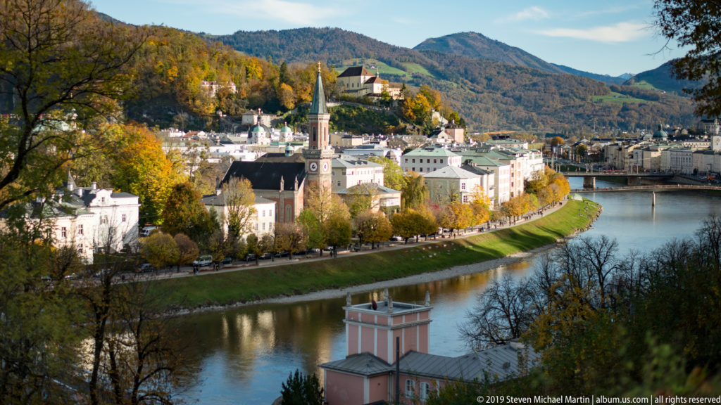 Elevated view of Salzburg’s historic center along the Salzach, with church towers, bridges, autumn trees, and wooded hills.