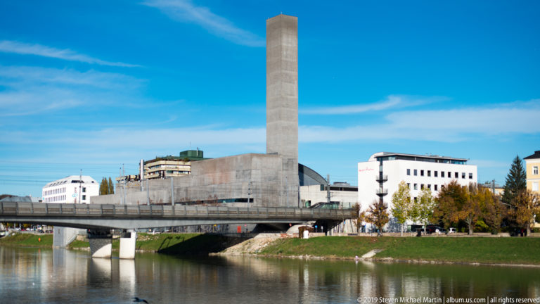 Concrete tower and modern riverside complex beside the Salzach, with bridge, grassy bank, and blue sky.