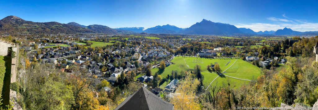 Elevated panoramic view over Salzburg neighborhoods and green fields toward a dramatic Alpine mountain range under blue sky.