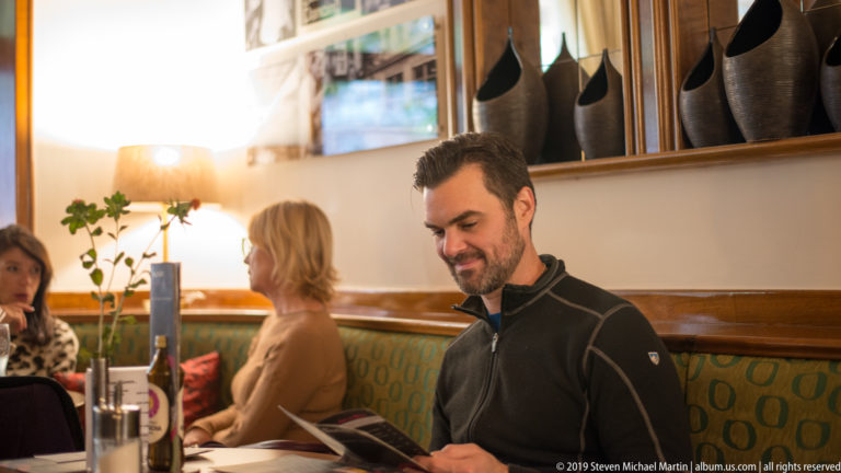 Man reading a menu at a restaurant table, with two diners beside him in a warmly lit Salzburg interior.