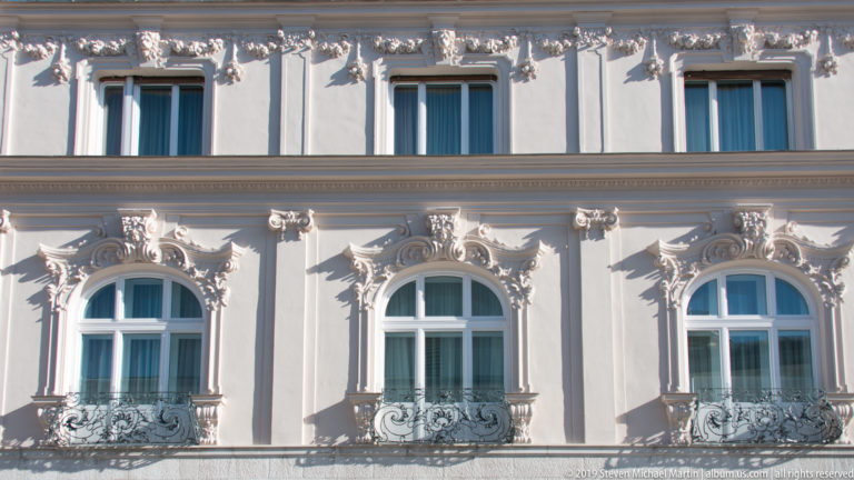 Close view of a pale historic façade with arched windows, ornate stucco surrounds, and small wrought-iron balconies.
