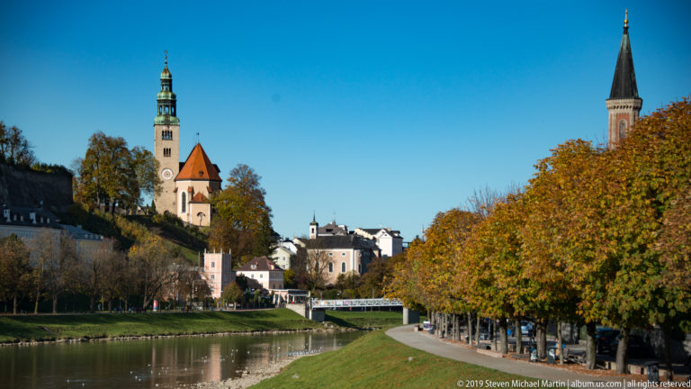 Müllner Kirche tower and another distant spire above a Salzach riverbank lined with dense golden autumn trees.
