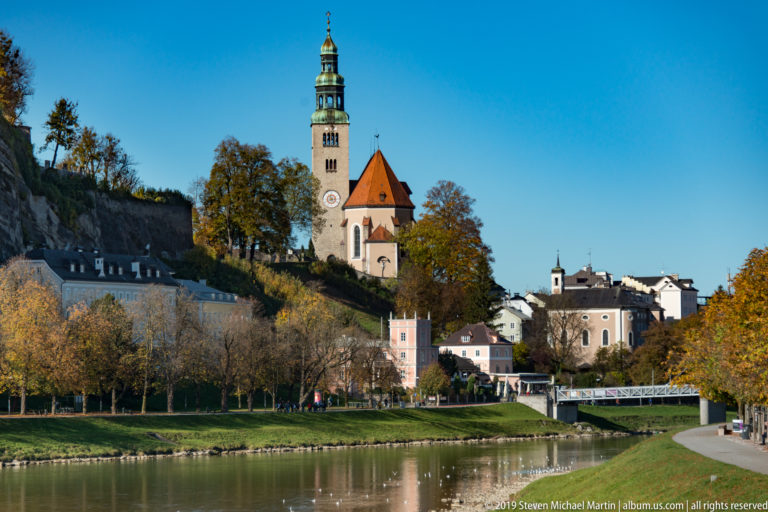 Müllner Kirche with green clock tower and red roof above the Salzach, framed by autumn trees and hillside.
