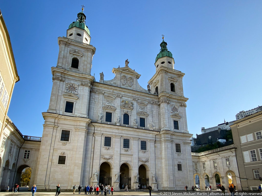 Salzburg Cathedral’s pale stone façade with twin green-domed towers, three entrance arches, sculptural ornament, and visitors crossing the square.