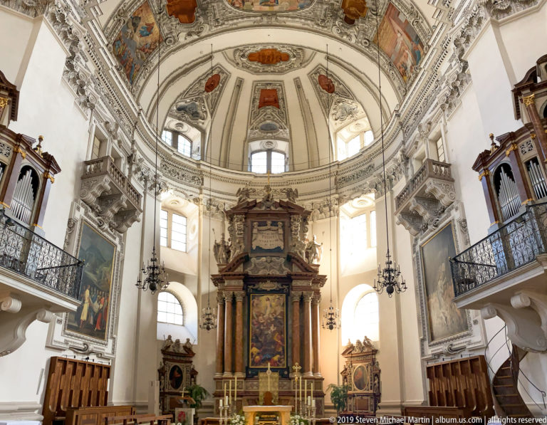High altar centered beneath decorated apse, with bright arched windows, organ galleries, chandeliers, side paintings, and choir furnishings.