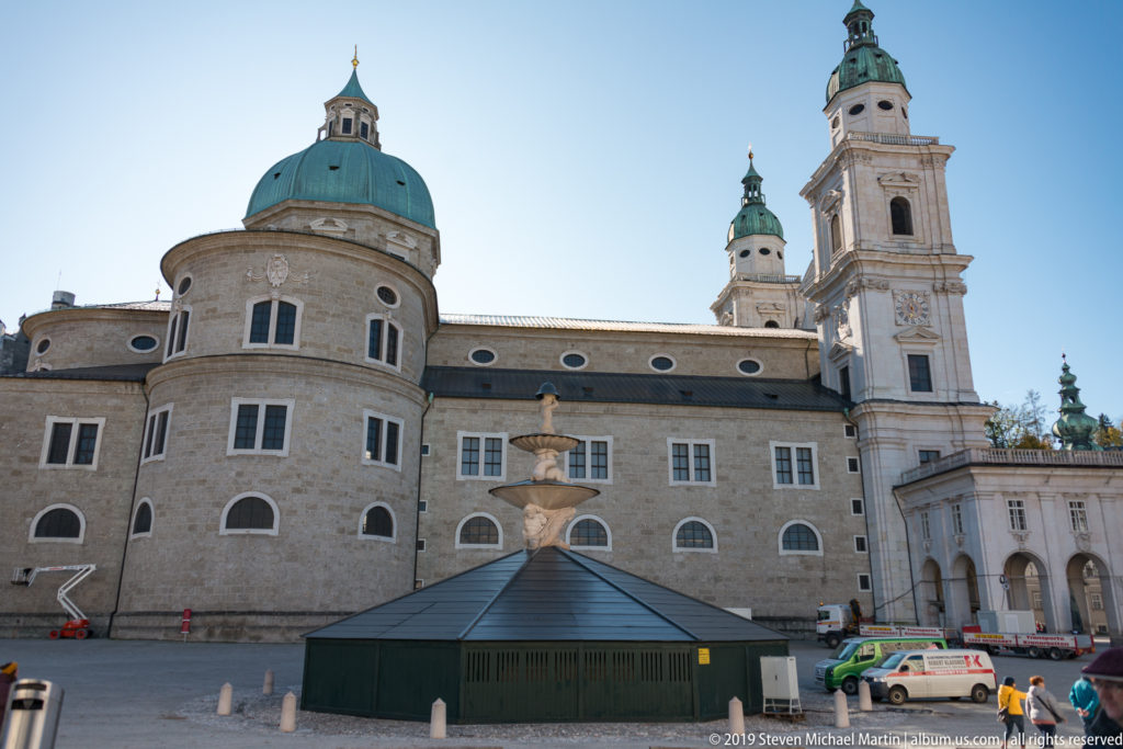 Rear elevation of Salzburg Cathedral with central green dome, right-hand tower, smaller lanterns, fountain, and dark temporary pavilion in the square.