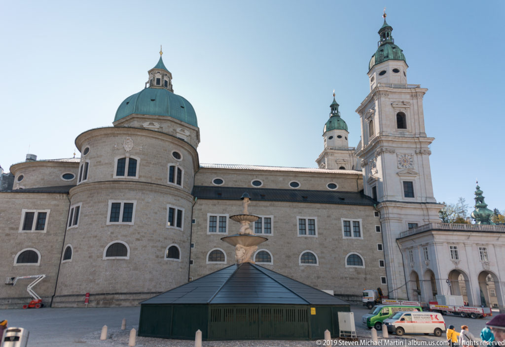 Wide rear-side view of Salzburg Cathedral with rounded masonry walls, green dome, tall tower, smaller roof lanterns, and square in foreground.