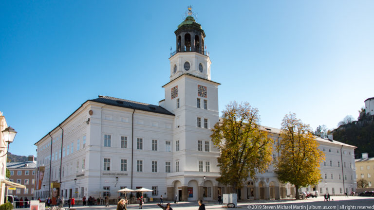 White historic civic building with arcaded ground floor and tall clock tower beside a square with pedestrians, café seating, and autumn trees.