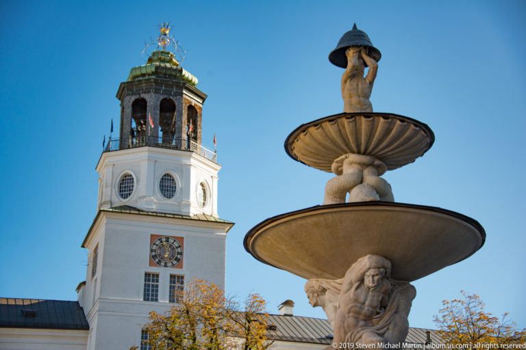 Tiered stone fountain with small crowning figure in front of a white clock tower, green roof, autumn foliage, and blue sky.