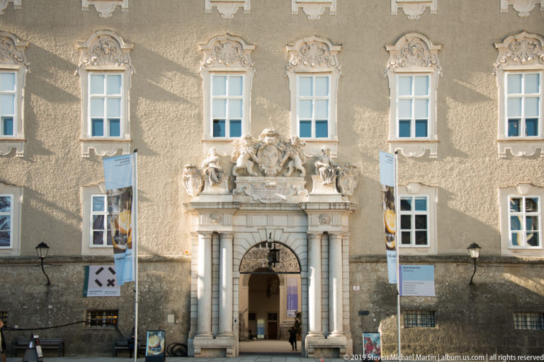 Historic façade with paired-column entrance, sculptural crest, arched doorway, decorated window surrounds, and vertical exhibition banners.