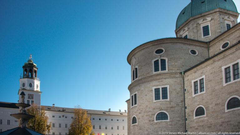 Rounded rear wall of Salzburg Cathedral beneath a green dome, with windows, a distant clock tower, and fountain element across the square.