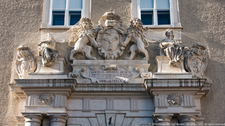 Ornate stone portal topped by a heraldic crest, sculpted lions, flanking figures, scrolling shields, and a central Latin inscription.