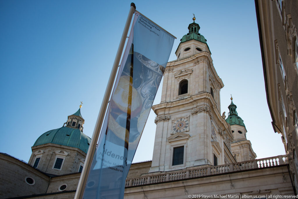 Cathedral tower and green dome rising above nearby buildings, partly obscured by a tall vertical banner in the foreground.