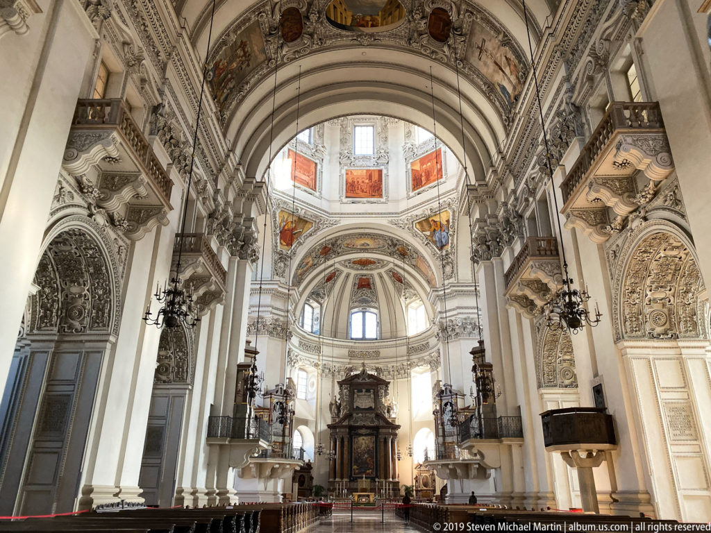 Vertical interior view of Salzburg Cathedral’s sanctuary beneath decorated arches and dome, with chandeliers and tall windows along the crossing.