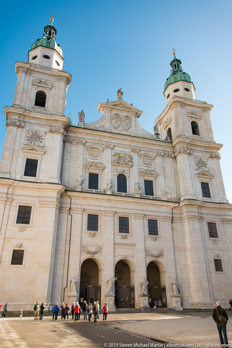 Full frontal view of Salzburg Cathedral façade with twin towers, central pediment, three arched portals, statues, and visitors across the base.