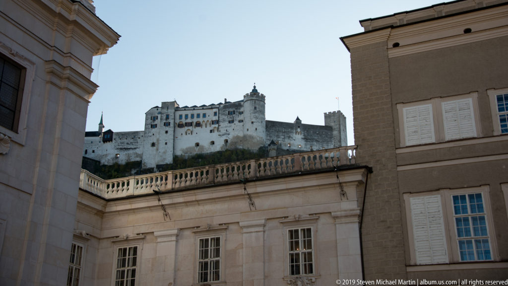 Hohensalzburg Fortress on the ridge above pale masonry buildings, balustrades, windows, and rooftops of the cathedral precinct.