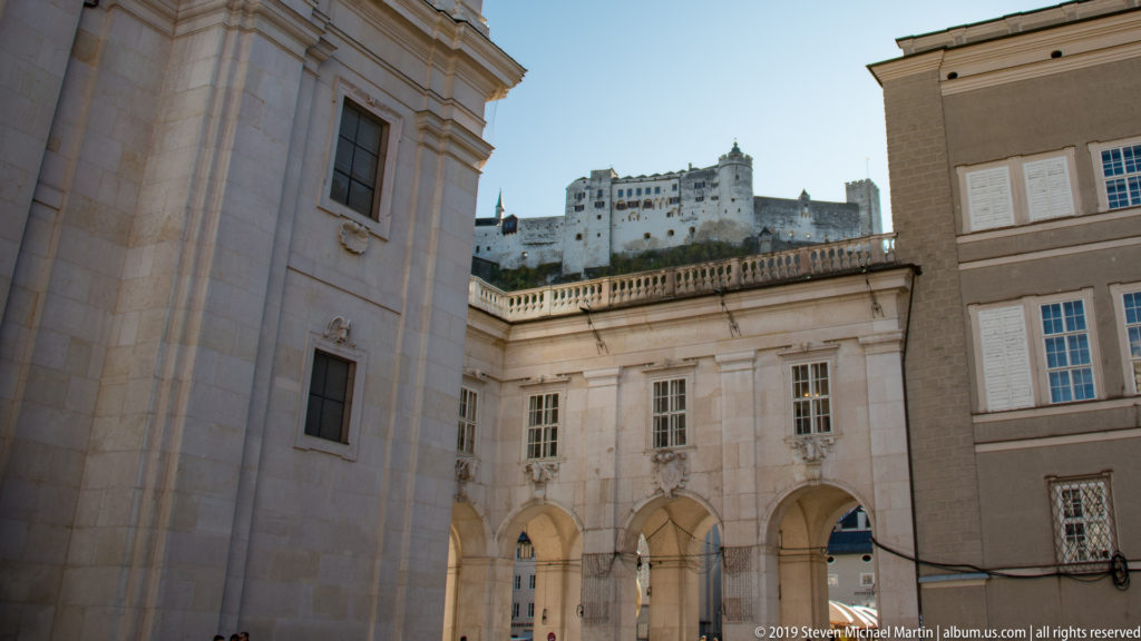 Arcaded courtyard buildings below Hohensalzburg Fortress, which rises on the ridge beyond under a clear blue sky.