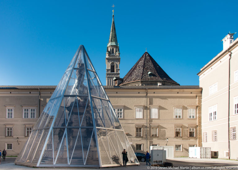 Glass pyramid pavilion in a paved square with historic façades, pitched roof, slender church tower, and a few pedestrians behind.