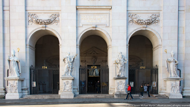 Three arched cathedral entrances with dark gates, monumental stone statues, inscriptions, relief ornament, and visitors at the façade.