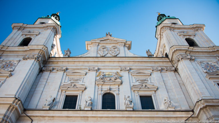 Low-angle view of Salzburg Cathedral’s pale stone façade with twin towers, sculptural niches, stacked windows, and central pediment.