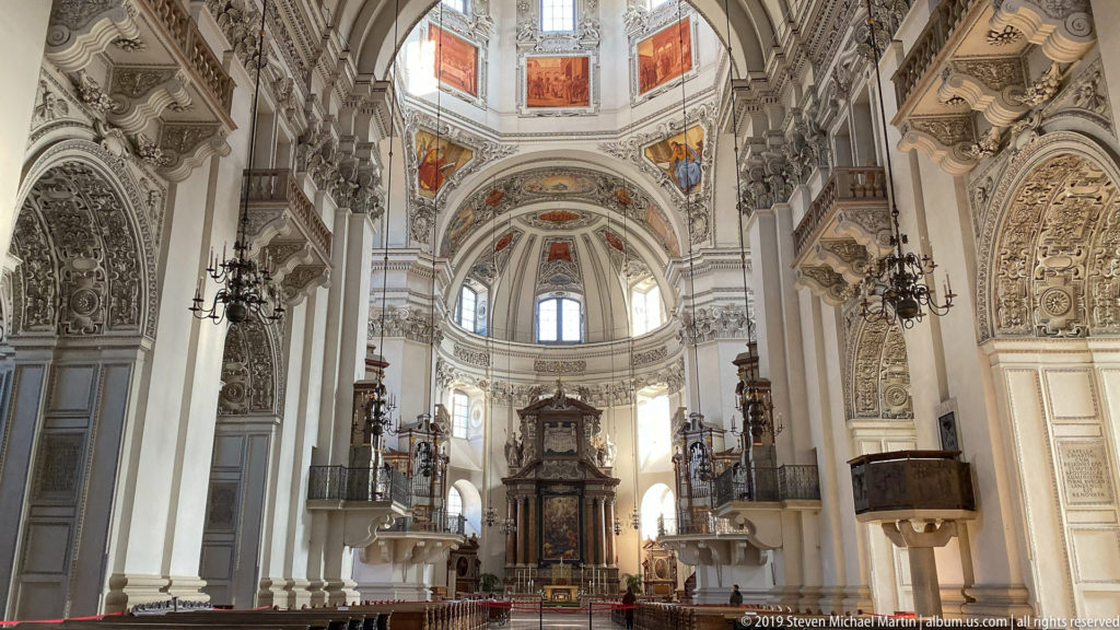 Axial view toward Salzburg Cathedral’s sanctuary and apse, framed by decorated piers, arches, chandeliers, windows, and projecting furnishings.