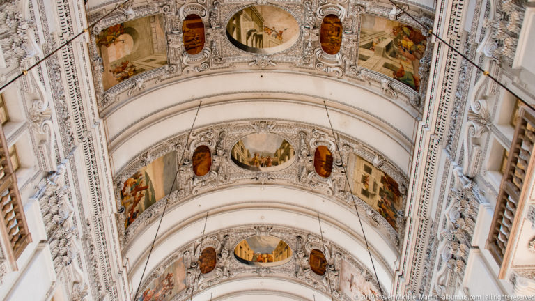 Oblique upward view along successive barrel-vault bays with paintings, oval medallions, ribs, and elaborate white stucco.