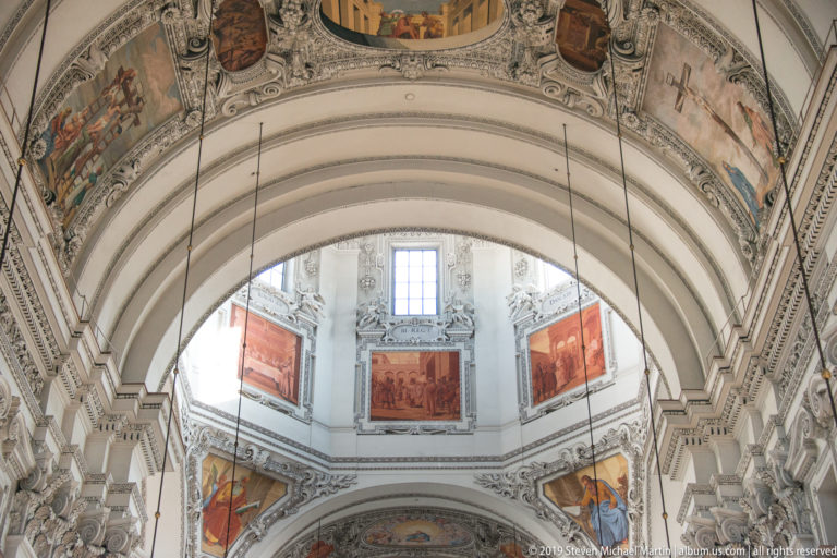 Upward view of broad nave arches, clerestory windows, painted panels, oval medallions, and white stucco decoration.