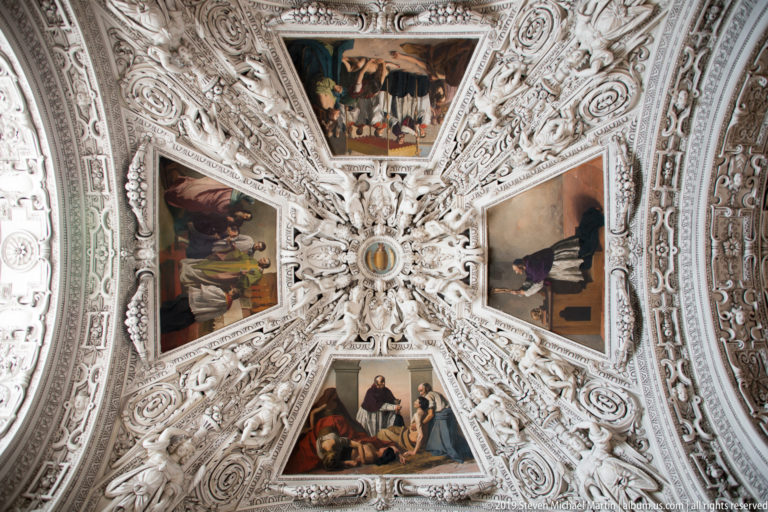 Four religious ceiling paintings arranged around a small circular center within dense white sculpted stucco ornament.