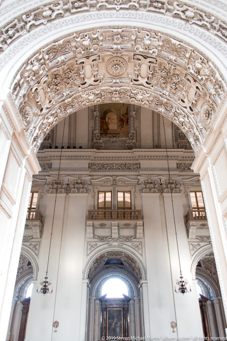 Low-angle interior view with ornate stucco arch, upper religious painting, paired windows, chandeliers, and a large arched doorway below.