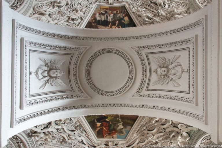 White vaulted ceiling bay with a circular central field, two sculpted angelic faces, molded borders, and adjacent painted decoration.