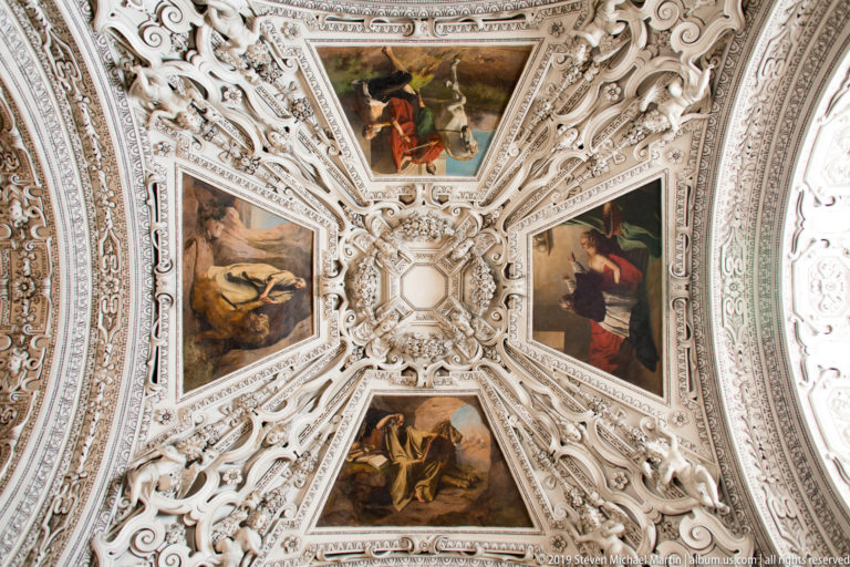 Ceiling detail with four religious paintings arranged around a central rosette amid dense white stucco scrollwork and sculptural ornament.