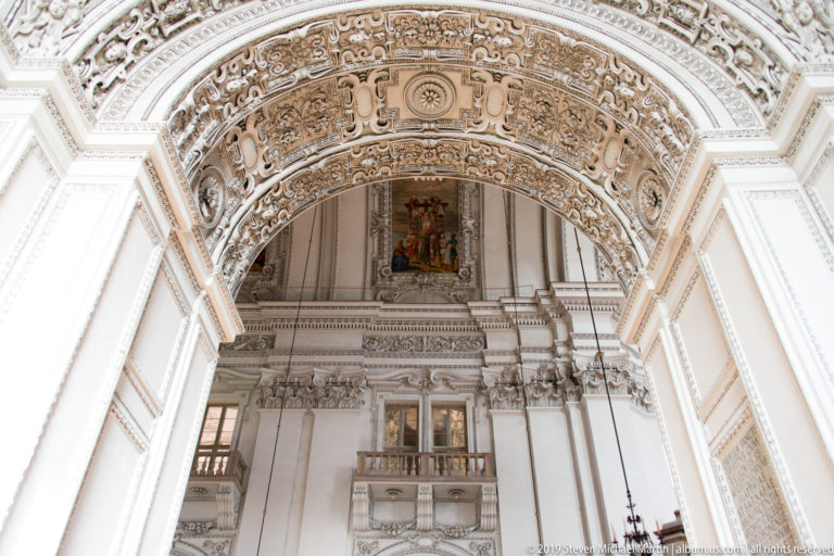 Upward view beneath an ornate stucco arch toward an upper wall bay with religious painting, paired openings, cornices, and pilasters.