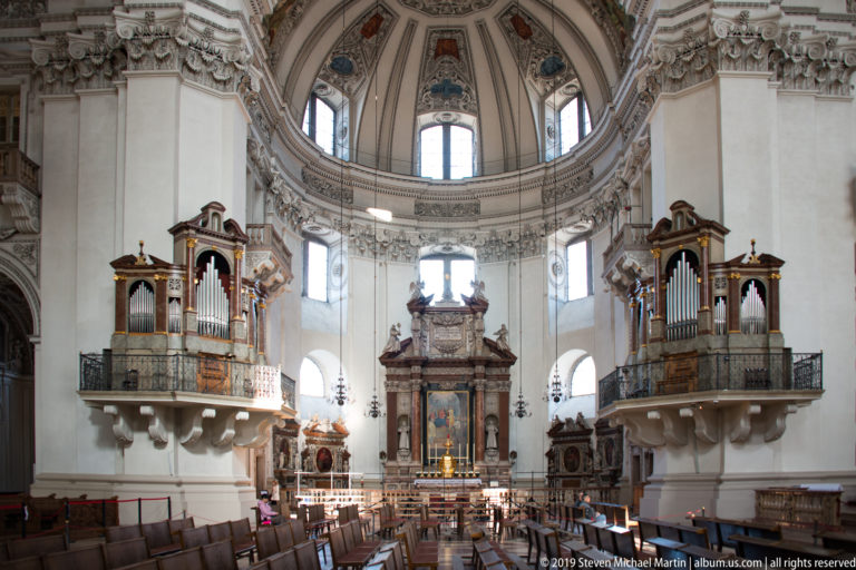 Symmetrical cathedral interior with high altar ahead, organ galleries on both sides, rows of chairs, arched windows, and dome above.