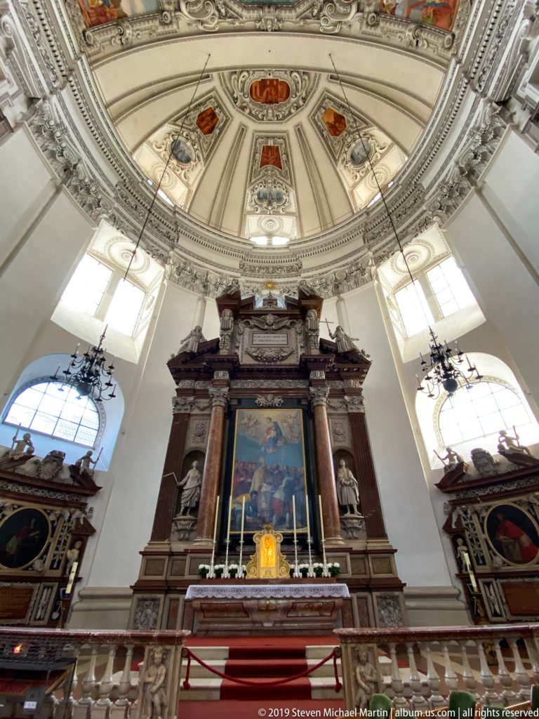 Monumental side altar with dark columns and large altarpiece beneath a richly decorated vault and tall windows in Salzburg Cathedral.