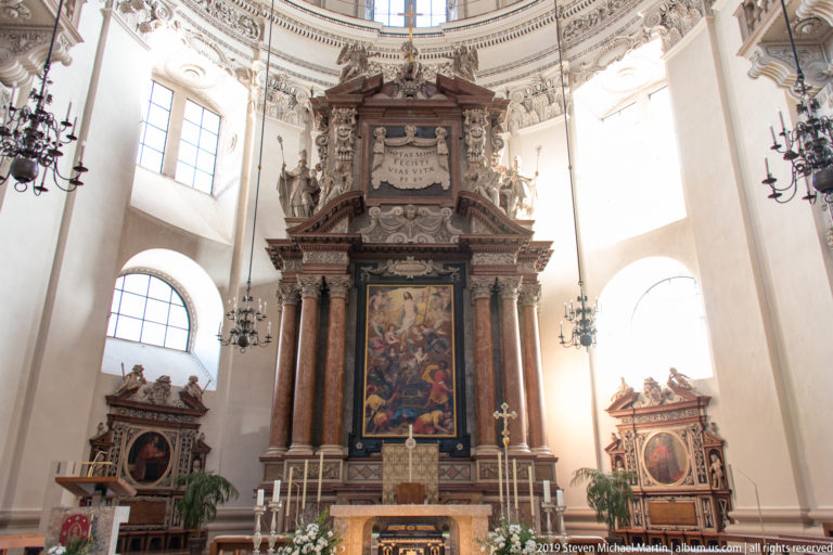 Monumental high altar with central painting and reddish marble columns, lit by tall arched windows on both sides.