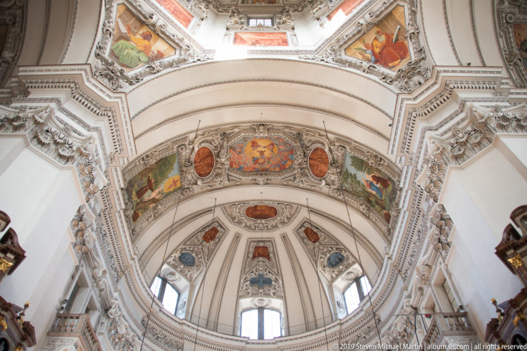 Large interior arch with painted panels, oval medallions, and white stucco beneath part of the cathedral dome.