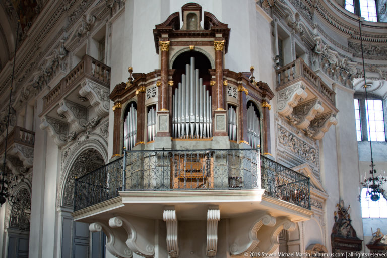 Pipe organ in a dark wooden case on a projecting gallery with iron railing and large white supports.