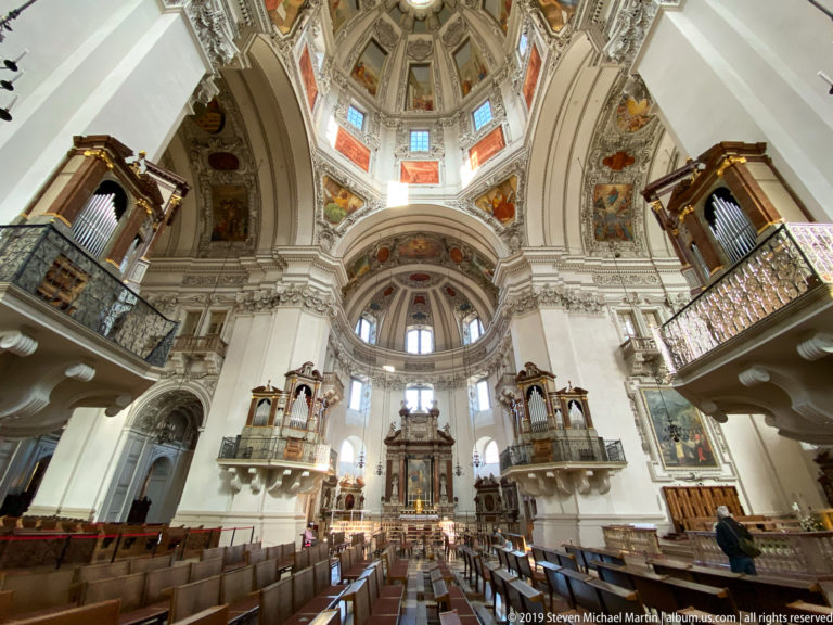 Wide interior view down Salzburg Cathedral’s nave toward the sanctuary, with pews, organ galleries, decorated arches, and the dome above.