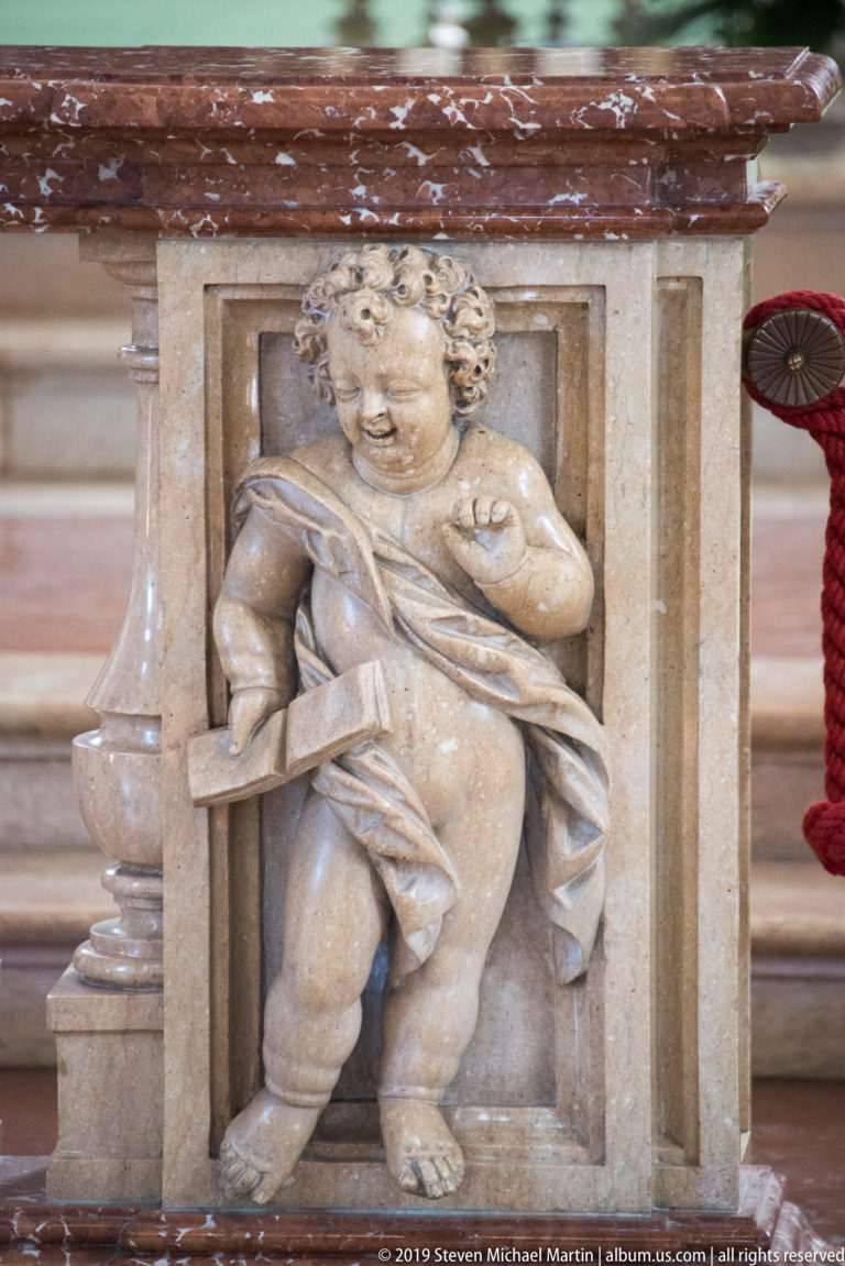 Small carved putto in high relief on a pale stone panel beneath a reddish marble ledge.