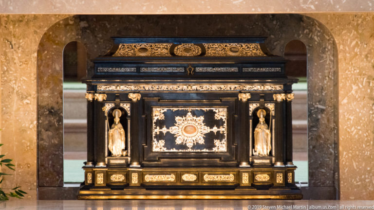Black-and-gold devotional cabinet with a radiant circular center, small flanking figures, columns, and gilded ornament.