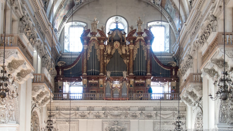 Large pipe organ in a carved dark wooden case above a gallery, framed by bright windows and white stucco.