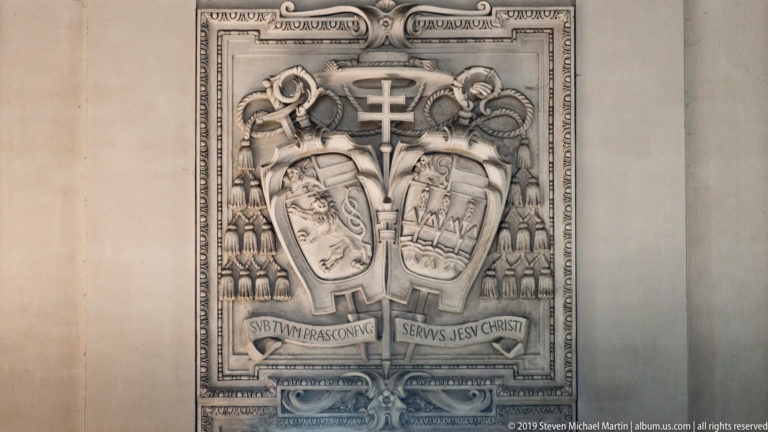 Pale carved heraldic relief with two shields, ecclesiastical tassels, central cross, and Latin motto ribbons.