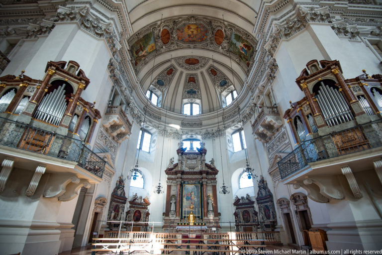 Wide interior view with high altar centered beneath the dome and large pipe organs on both side galleries.